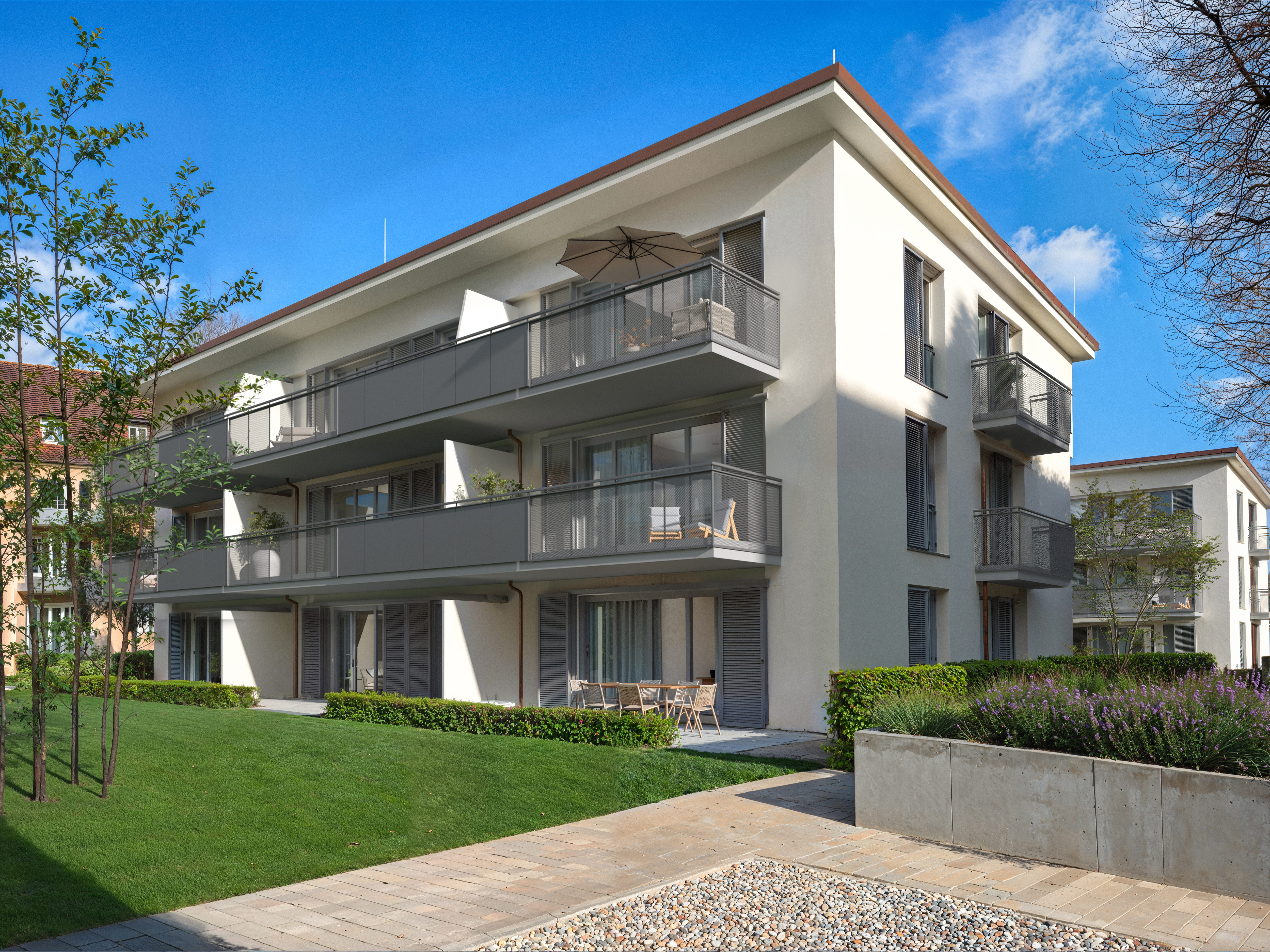 Fotorealistisches Render des fertiggestellten und voll möblierten Mehrfamilienhauses. Das dreistöckige Gebäude mit heller Putzfassade präsentiert sich mit dunkelgrauen Balkongittern. Die Erdgeschoss-Terrasse ist einladend mit einer Essgruppe möbliert, die darüber liegenden Balkone zeigen Lounge-Möbel und einen Sonnenschirm. Ein gepflegter grüner Rasen, Hecken und eine Betonmauer mit Lavendelpflanzung im Vordergrund unterstreichen die hochwertige Außenanlage.